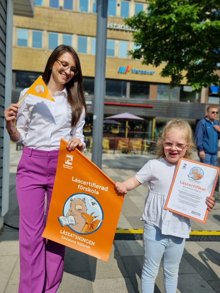 En vuxen och ett barn med den orange flaggan och diplomet för läscertifierad förskola.